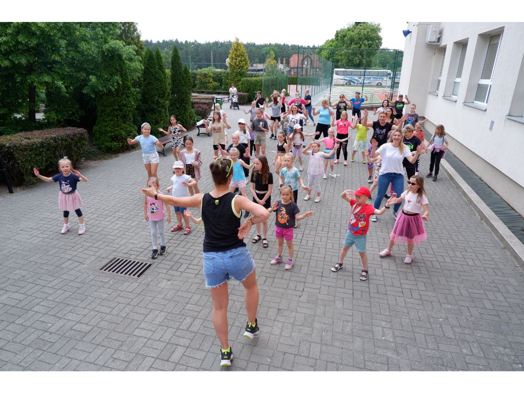 Grupa dzieci i dorosłych kobiet tańczących przed budynkiem.