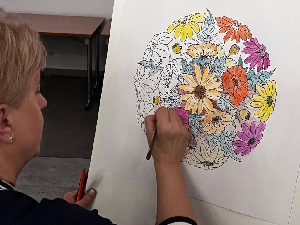 Kobieta siedząca przed sztalugą, koloruje kredką kwiaty na białej kartce.